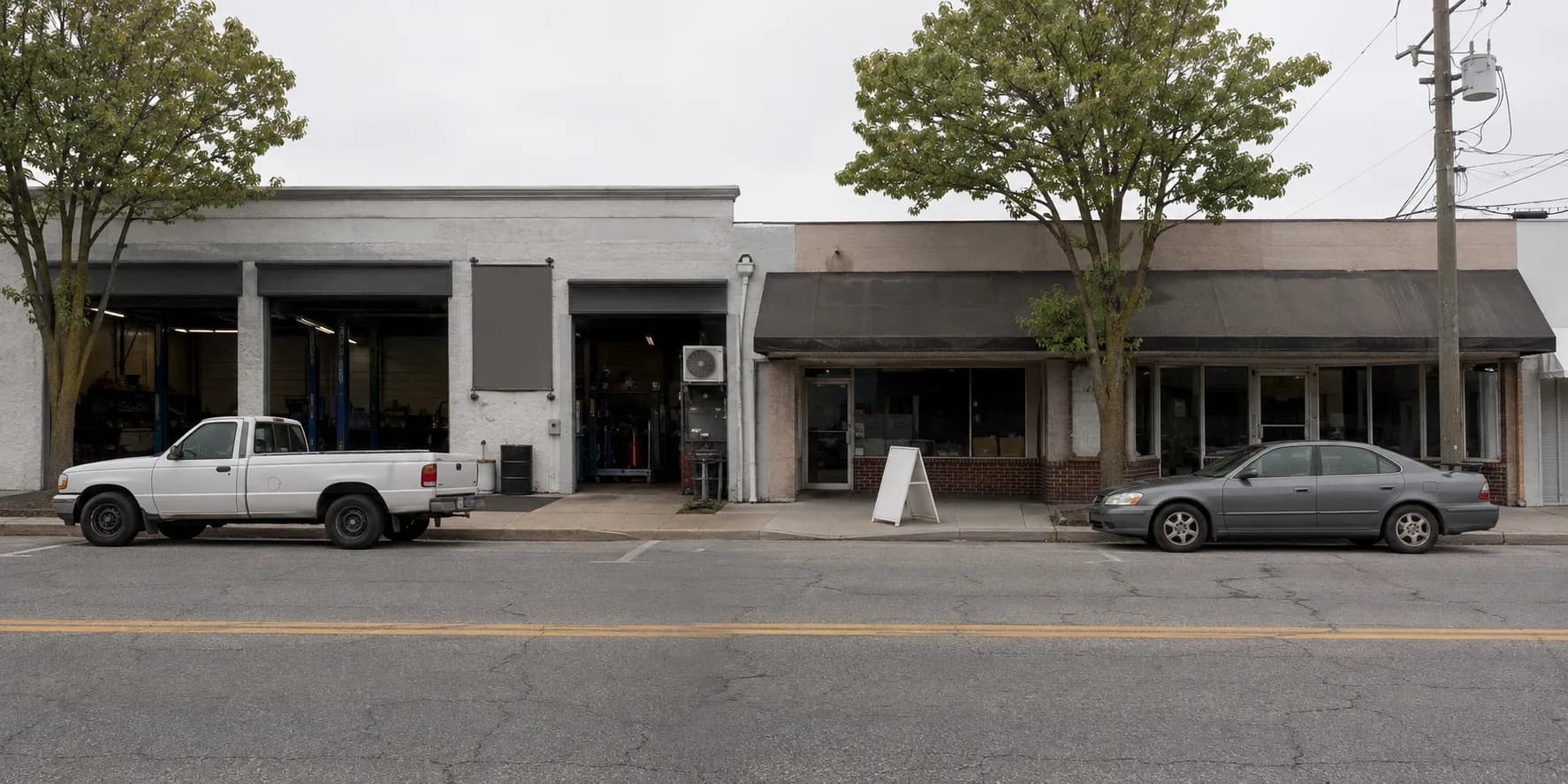 A block of independent businesses on an American commercial street on a weekday morning: an auto repair shop with its bay doors rolled up, a storefront under a faded awning next door, a pickup and a sedan parked at the kerb